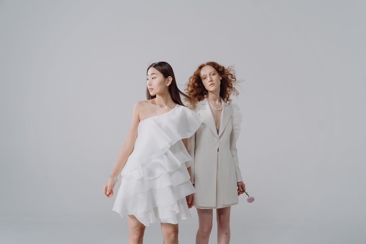 Menu Two women in stylish white dresses posing gracefully in a studio setting against a light background.